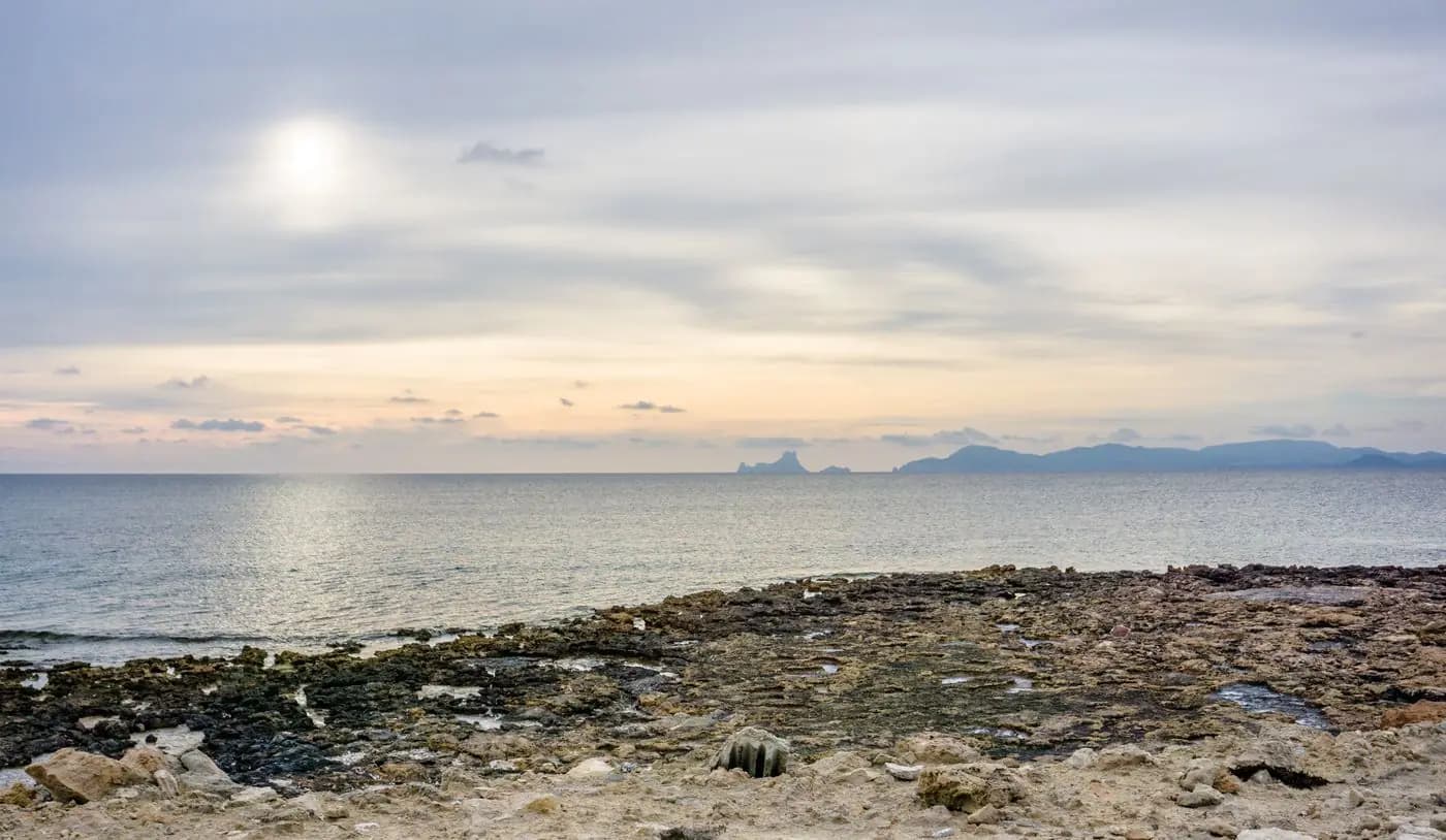Rotsachtige kust bij zonsondergang, met het eilandje Es Vedrà aan de horizon